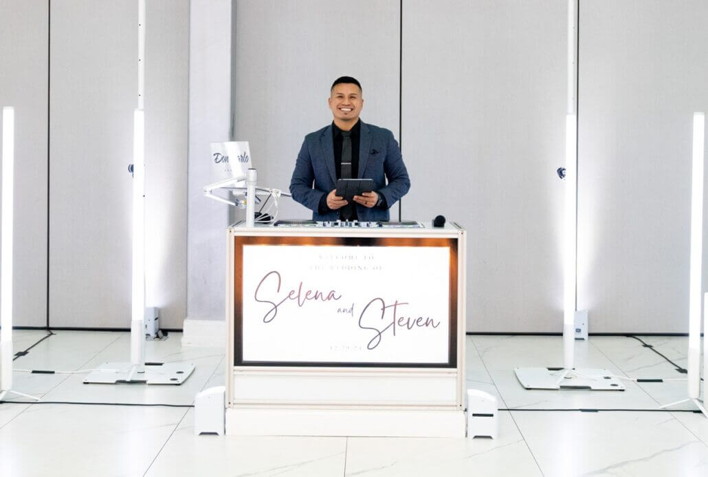 A man stands behind a DJ booth with a sign reading "Selena and Steven." The area is lit by vertical lights and has a minimal, modern background.
