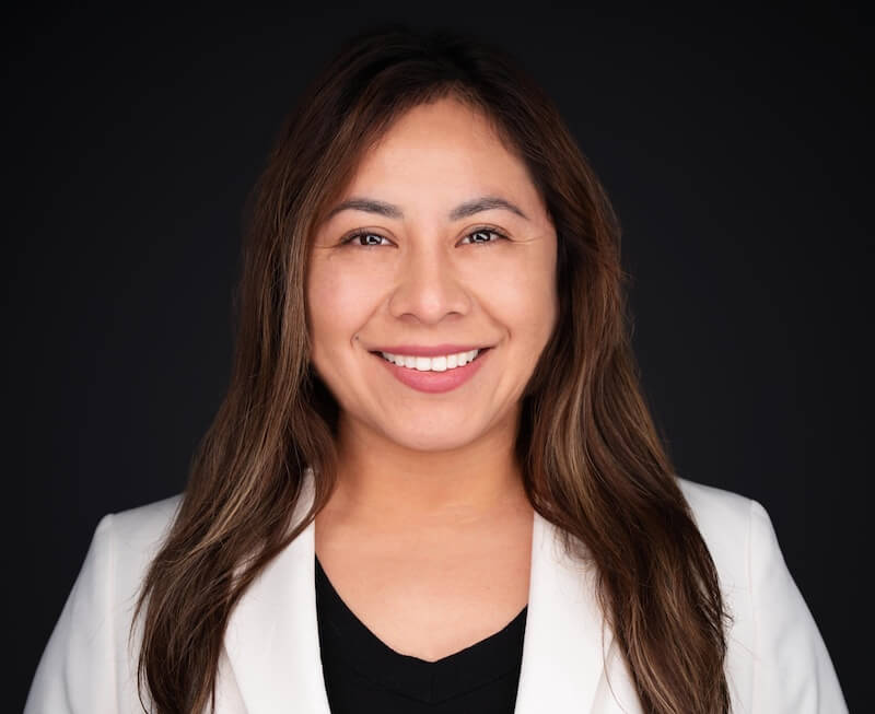 Woman with long brown hair smiles at the camera, wearing a white blazer over a black top, with a dark background.