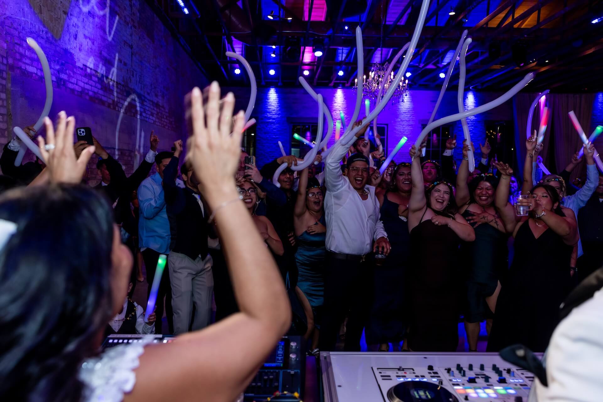 A DJ plays music while a crowd of people on a dance floor wave long white balloons and raise their hands under blue and purple lights.