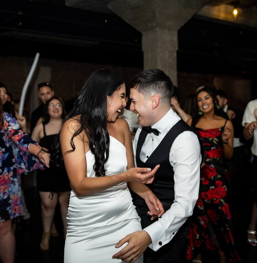 A couple in formal attire laughs and dances closely together at a lively indoor event, surrounded by other dancing guests.