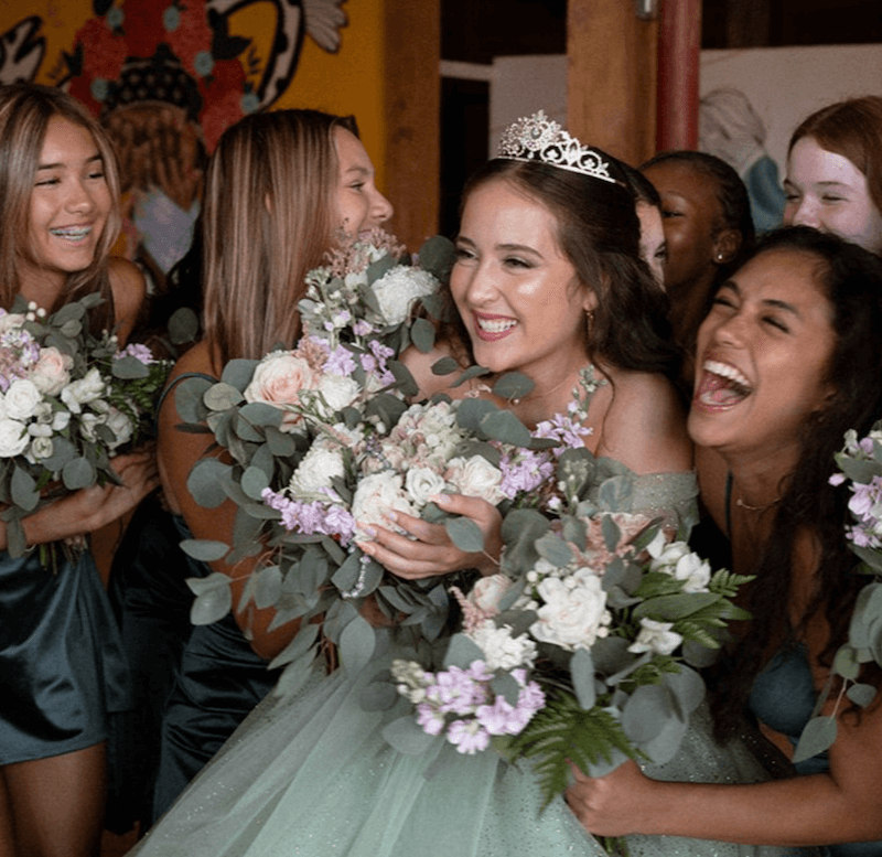 A group of young women in formal dresses, holding flower bouquets, surround and celebrate a girl wearing a tiara and a light green dress.