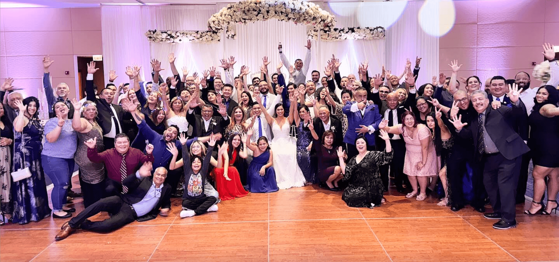 A large group of people in formal attire pose together on a dance floor at a wedding reception, smiling and raising their hands toward the camera.