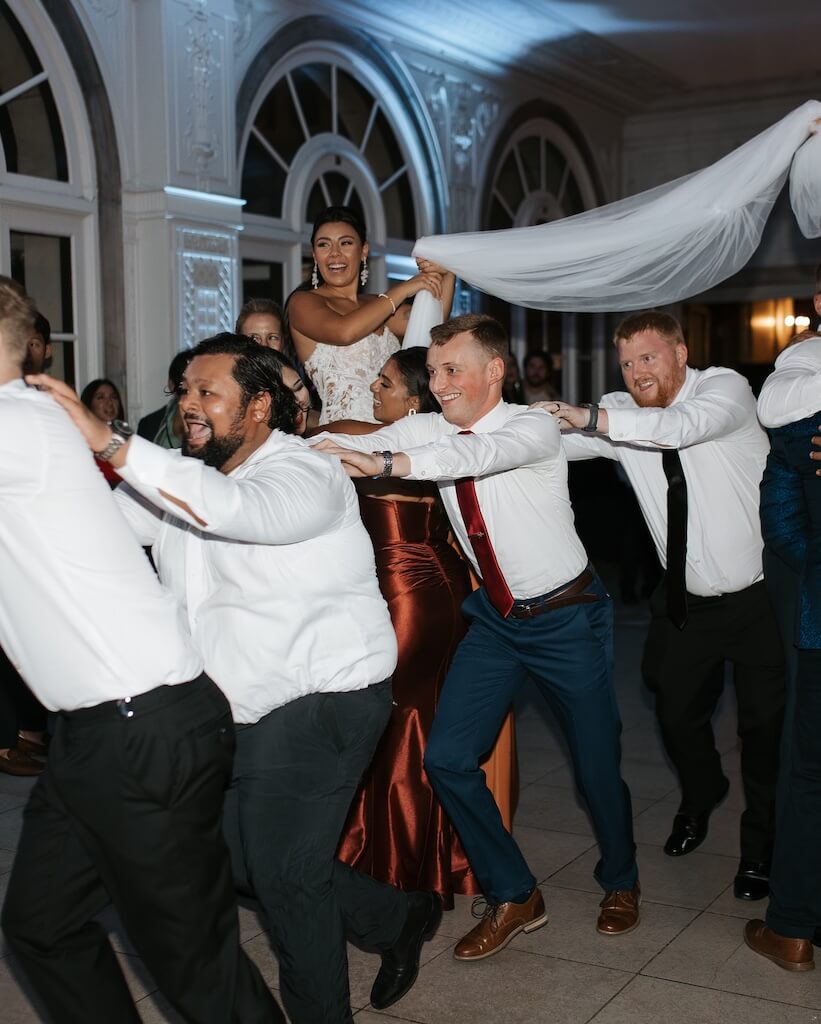 A bride is lifted on a chair while guests in formal attire dance in a conga line at an indoor wedding reception.