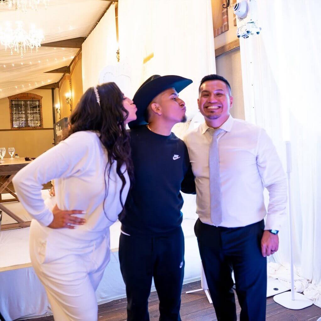 Three people pose indoors; a woman in white and a man in a black Nike tracksuit pretend to kiss each other, while another man in a white shirt and tie stands smiling beside them.