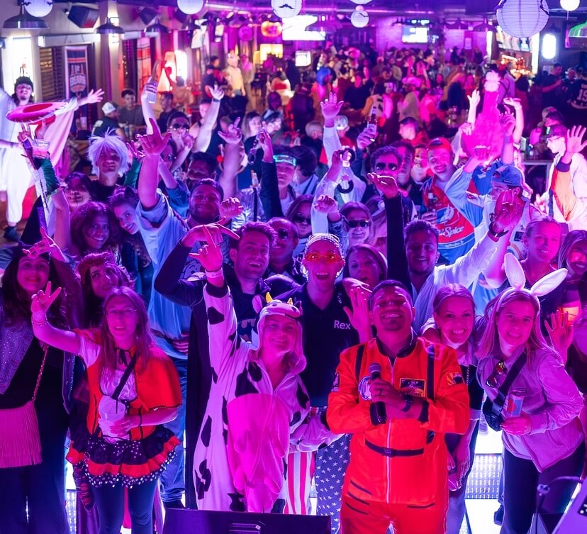 A large group of people in colorful costumes pose and smile on a stage in a lively, crowded indoor venue with lights and decorations.