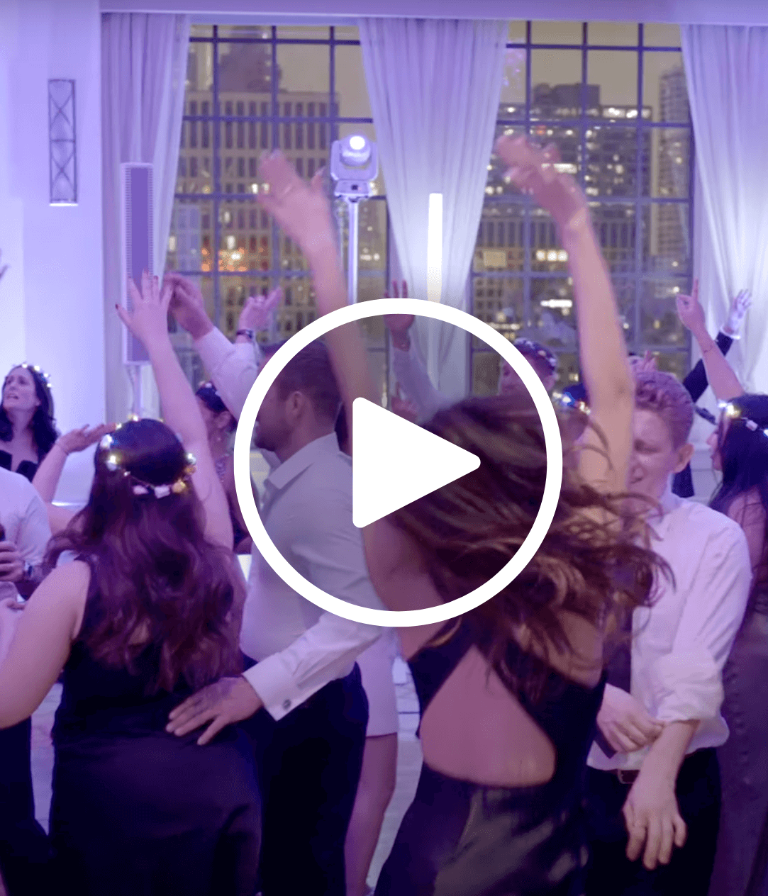 A group of people in formal attire dance with raised arms at an indoor event with large windows showing a cityscape at dusk.