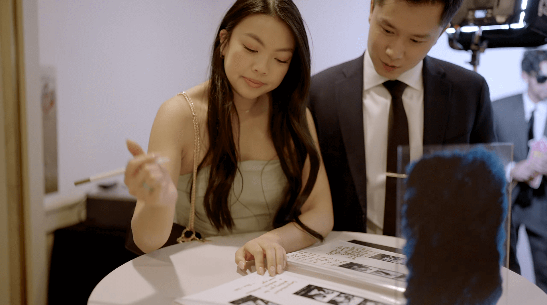 A woman and a man in formal attire look at and write in a guest book on a round table at an indoor event.