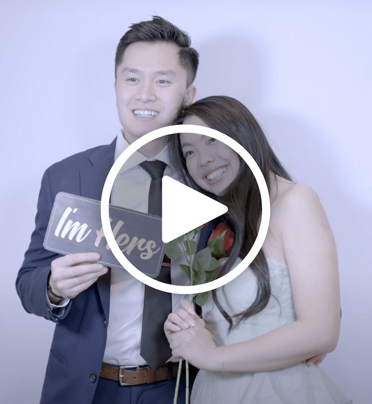 A man in a suit holds a sign that reads "I'm Hers" while a woman in a strapless dress hugs him and holds a red rose.