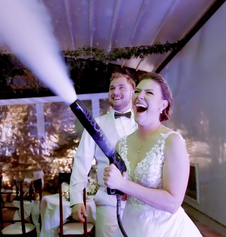 A bride in a white dress laughs while using a fog machine, with a groom in a white suit smiling beside her at a wedding reception.