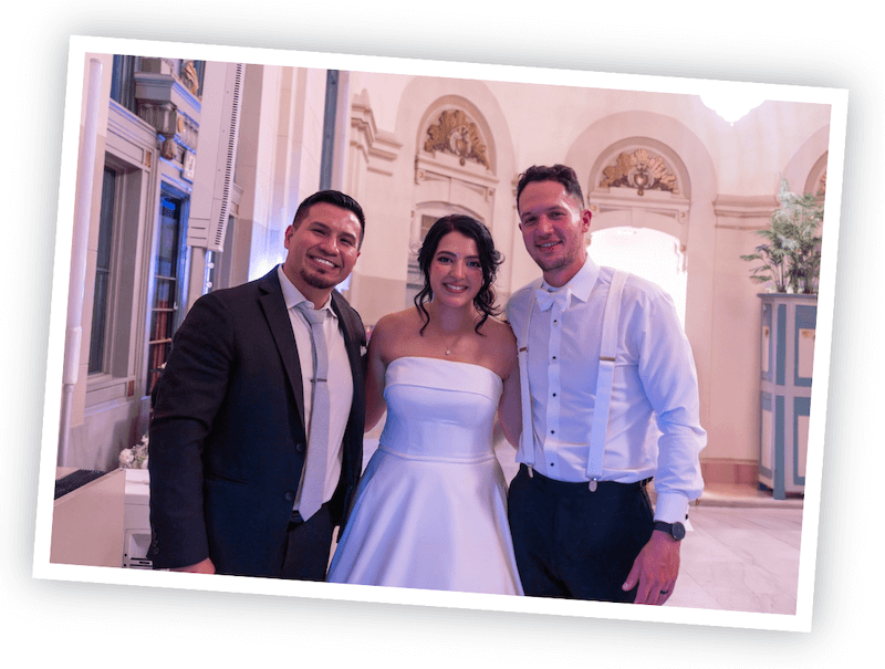 Three people posing indoors at a formal event; the woman in the center wears a strapless white dress, flanked by two men in dress shirts and pants, one in a suit and tie.