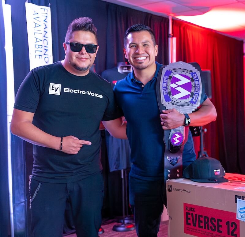 Two men stand indoors; one wears sunglasses and an Electro-Voice shirt while the other holds a large championship belt. They pose next to a box labeled "Everse 12" and a branded cap.