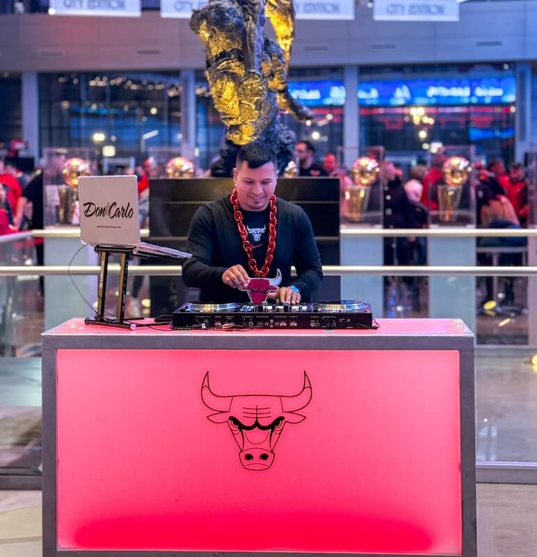 A DJ stands behind a lit DJ booth with a Chicago Bulls logo, mixing music at an indoor event with people and trophies in the background.