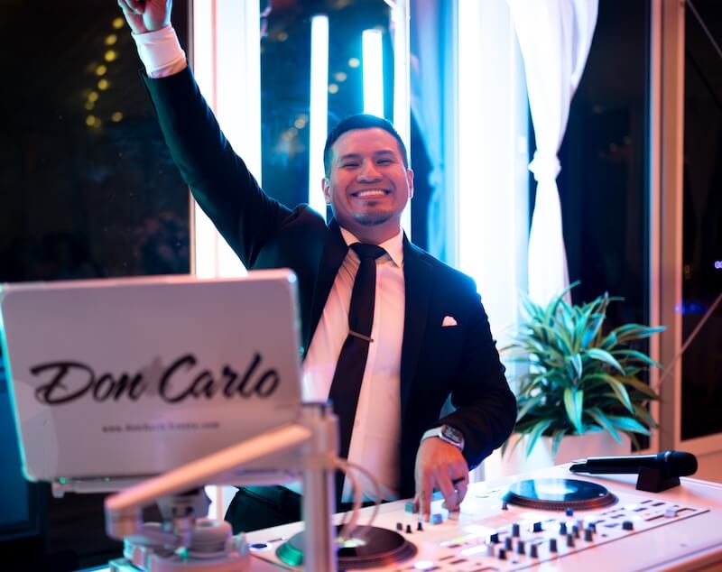 A man in a suit smiles and raises one arm while standing behind a DJ booth labeled "Don Carlo" with DJ equipment and bright lights in the background.