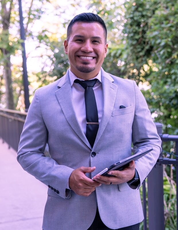 A man in a light gray suit holds a tablet and smiles while standing outdoors near a railing with trees in the background.