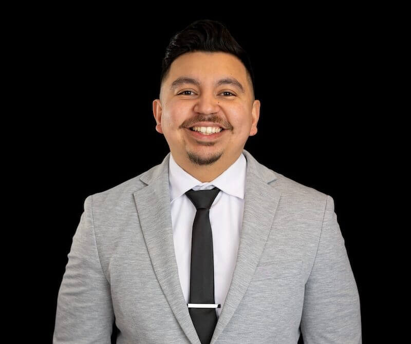 A man in a light gray suit, white shirt, and black tie stands smiling against a plain black background.