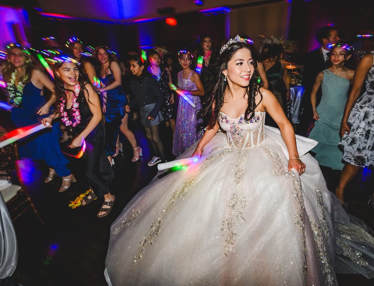 A woman in a formal gown and tiara, likely at a celebration, smiles while walking among a group of people dancing with glow sticks in a dimly lit room.