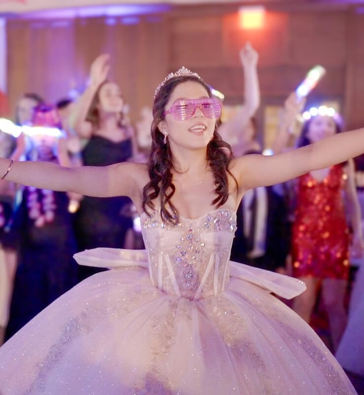 A young woman in a sparkling ball gown and pink sunglasses extends her arms while dancing at a party; people dance in the background.