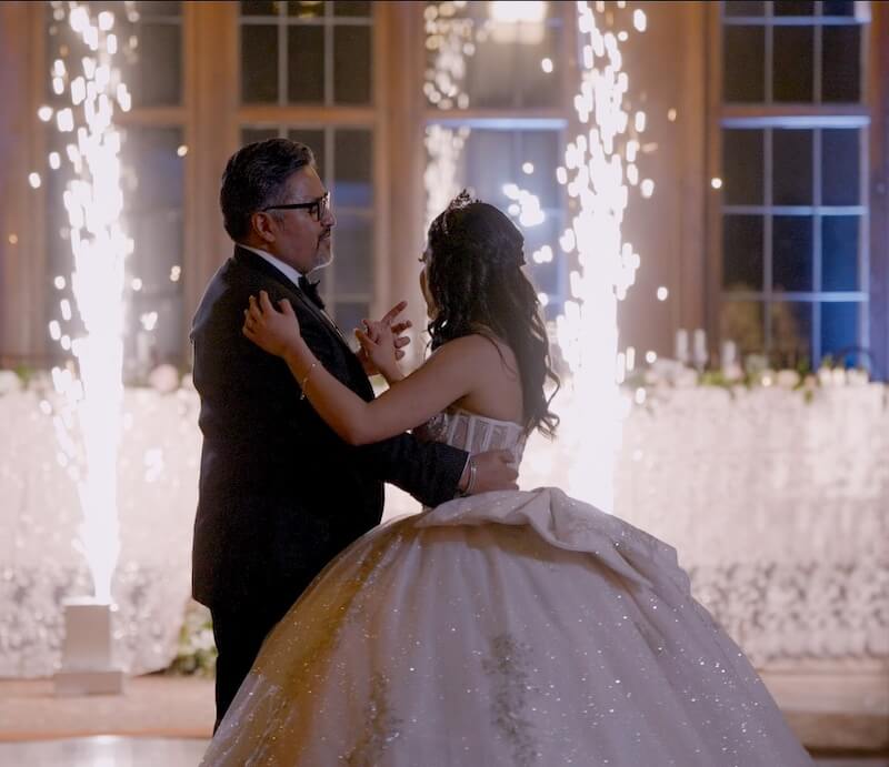 A man and a young woman in a formal gown dance together indoors, with bright fireworks in the background.