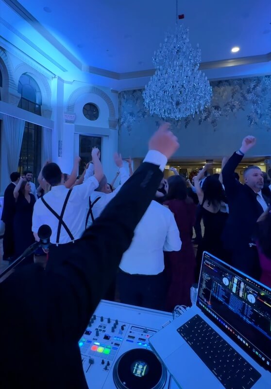 A group of people dressed formally dance with raised arms under a large chandelier, while a DJ plays music from a laptop and mixer in a blue-lit event hall.
