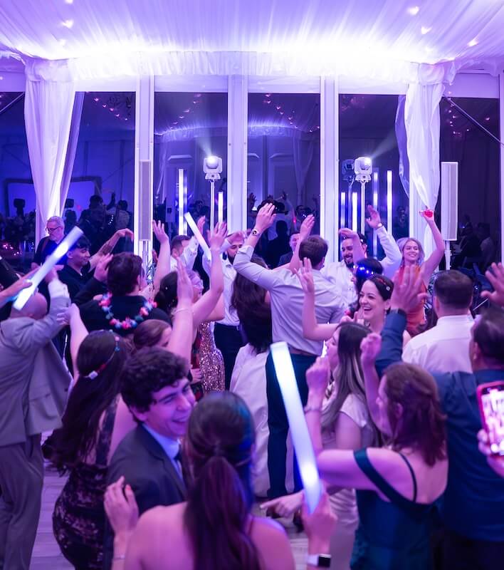 A group of people in formal attire dance and celebrate under a white tent with purple lighting, some holding glowing sticks.
