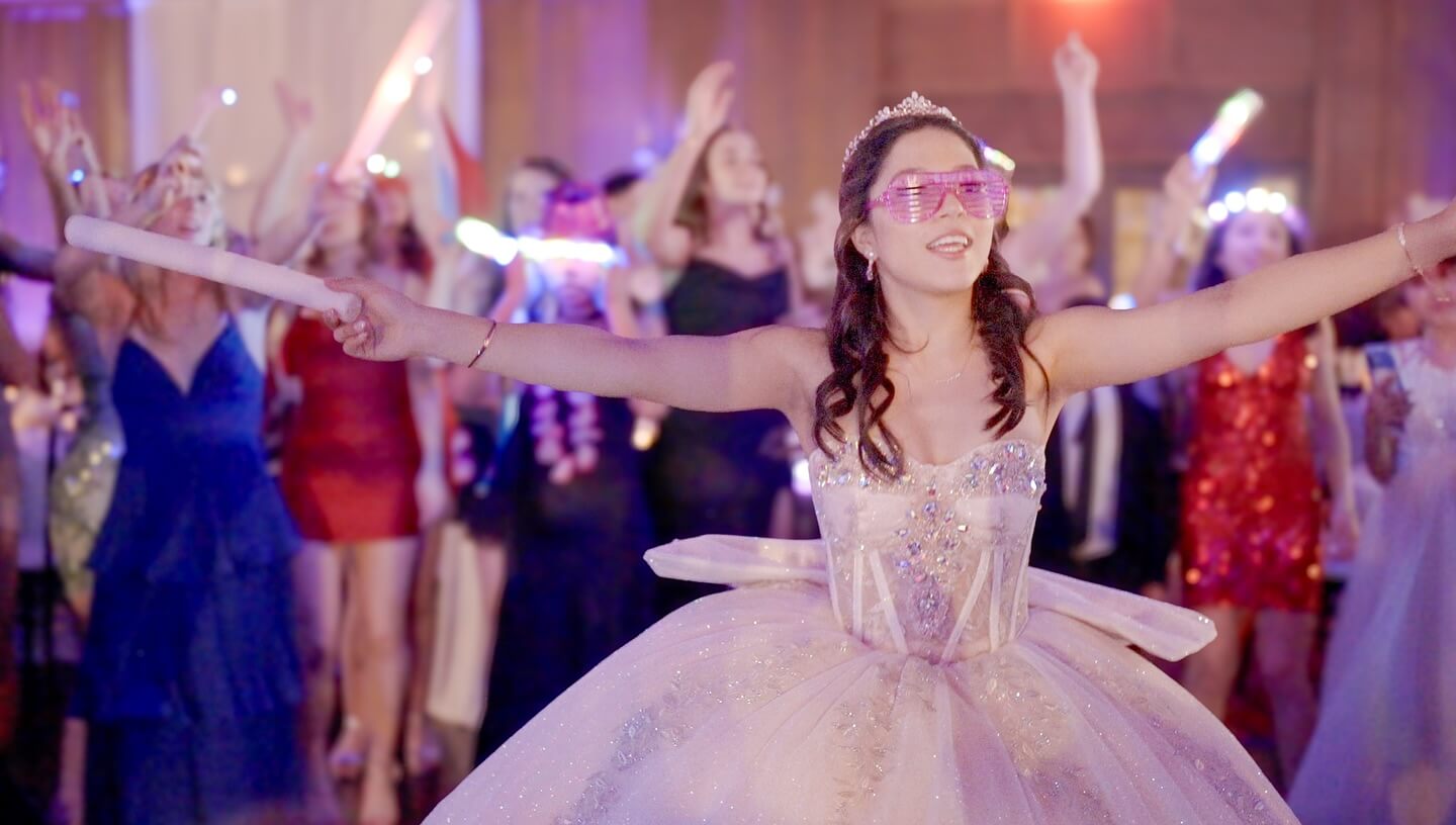 A young woman in a formal, sparkling gown and pink glasses stands with arms outstretched at a party, as a Cicero IL Bilingual DJ energizes the dancing crowd amid colorful lights in the background.