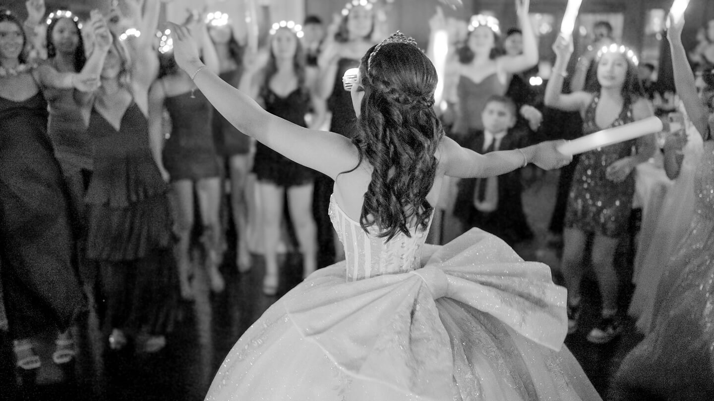 A girl in a formal gown and tiara stands with arms raised, facing a cheering group of people at a festive event with music by a Cicero IL Bilingual DJ.