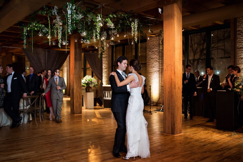 A bride and groom share their first dance in a warmly lit venue with wooden floors, surrounded by wedding guests and hanging greenery—perfect inspiration for couples exploring Greater Chicago Wedding Venues.