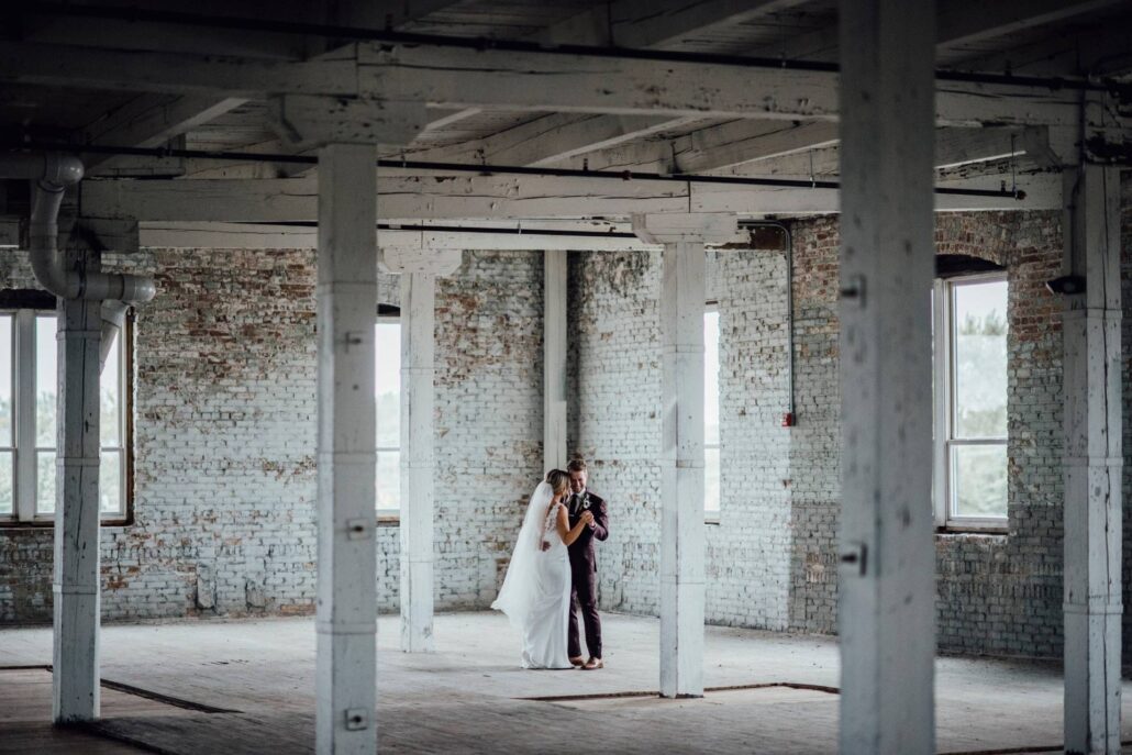 A bride and groom stand together in the middle of an empty, industrial-style room with exposed brick walls and large windows—a stunning example of Greater Chicago Wedding Venues.
