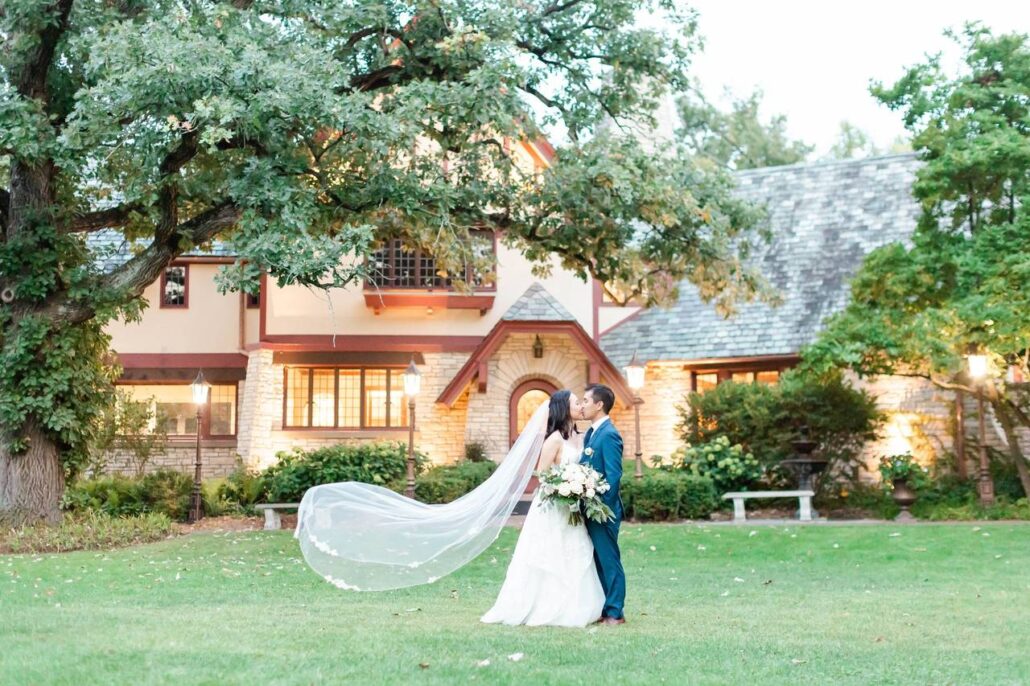 A bride and groom stand close together outside a large house at one of the stunning Greater Chicago Wedding Venues, with the bride’s veil flowing behind her and trees surrounding the scene.