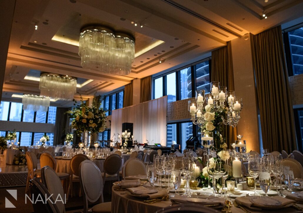 Elegant banquet hall set for a formal event with round tables, white chairs, floral centerpieces, and candlelit chandeliers, with city buildings visible through large windows—perfect for those seeking top Greater Chicago Wedding Venues.