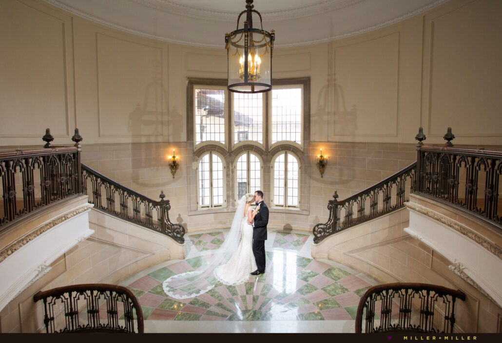 A bride and groom stand together at the center of a grand staircase in an elegant, well-lit hall with large windows and ornate railings—one of the premier Greater Chicago Wedding Venues.