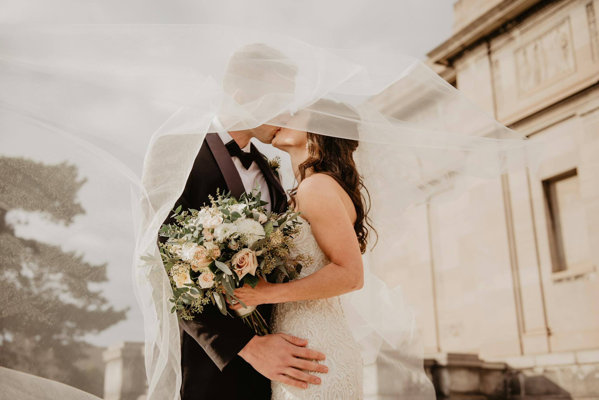 A bride and groom embrace and kiss under a flowing veil outdoors at one of the elegant Greater Chicago Wedding Venues, with the bride holding a bouquet and a stone building visible in the background.