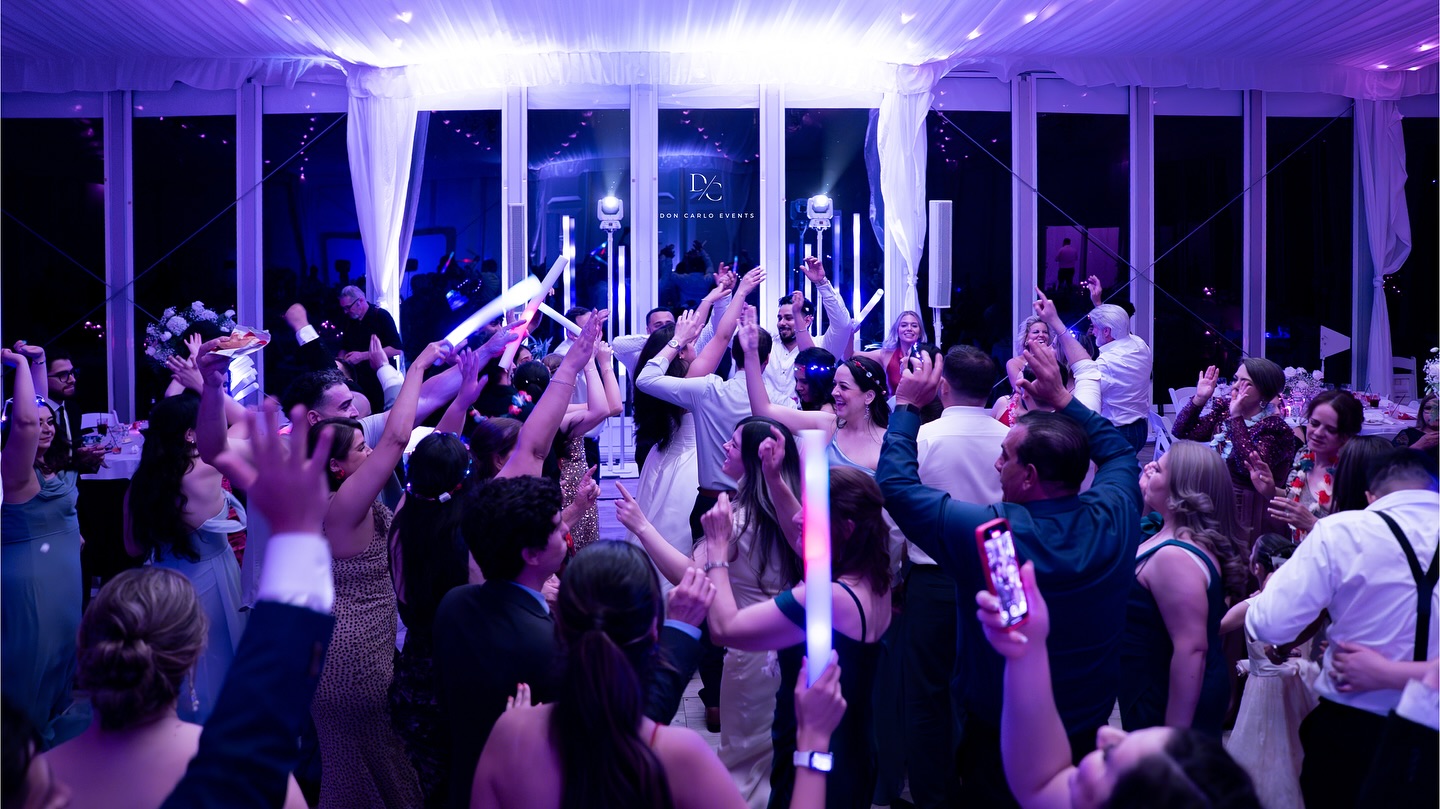 A large group of people dance and celebrate under purple lighting at an indoor event, many holding up glowing sticks.