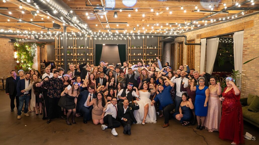 A large group of people in formal attire pose together indoors under string lights at a wedding reception, with the bride and groom in the center, showing how a bilingual DJ blends cultures to create an unforgettable celebration.